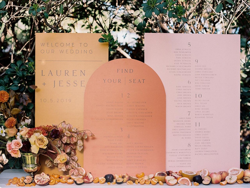 Romantic Boho Desert Pastel Wedding Ideas | A geometric trio of wedding signs in pink, dusty orange and terracotta tones styled with an autumnal floral arrangement in a vintage silver vase and scattered oranges, grapefruit, apples, plums and pomegranates. The first dusty orange rectangular sign reads, "Welcome to our wedding" with the couple's name and wedding date. The central terracotta arch shaped sign reads, "Find your seat" and lists table numbers and guest names. This seating chart is continued onto the third pink rectangular sign. | Photography: Amanda Crean via Style Me Pretty