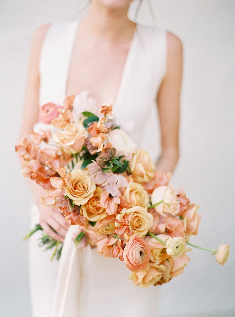 Romantic Boho Desert Pastel Wedding Ideas | A modern romantic asymmetrical wedding bouquet featuring pale yellow roses, peach ranunculus, salmon pink bougainvillea and blush hellebores tied with white flowing ribbon. No greenery is used to make this warm colour palette pop. | Photography: Ashley Ludaescher