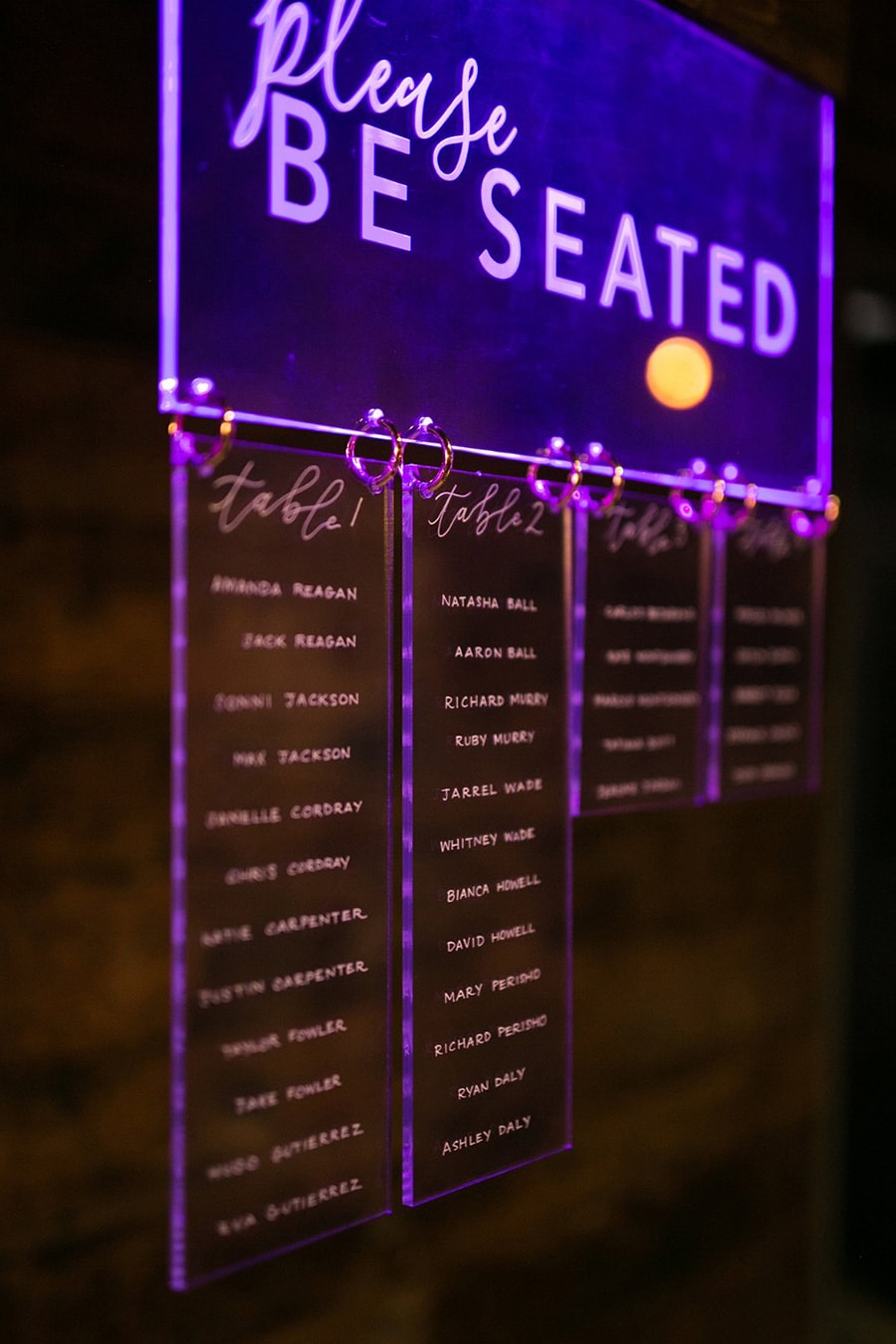 Electric Disco Wedding Ideas |  An acrylic wedding reception seating chart illuminated by purple lighting which reads "Please Be Seated". Table numbers and guest names are displayed on individual acrylic rectangles hang attached by metal rings | Photography: Amanda Grace Photography via Wedding Chicks