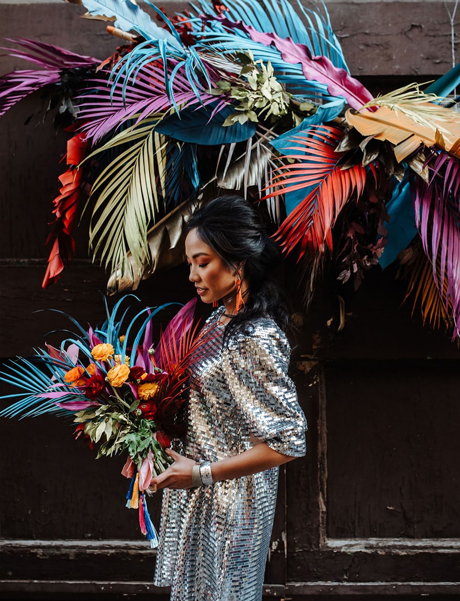Electric Disco Wedding Ideas |  Suspended wedding ceremony backdrop featuring a cluster of palm fronds and tropical greenery spray painted in bright neon colours. A bride stands in front of the installation wearing a silver mirror ball inspired wedding dress and holding a bright tropical wedding bouquet to match the backdrop | Photography: Silvia Galora Photography via Green Wedding Shoes