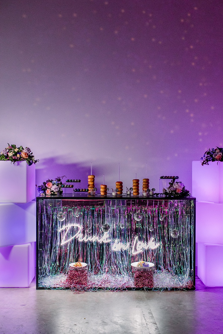 Electric Disco Wedding Ideas |  A disco inspired wedding dessert table featuring silver foil curtain, a "Drunk in Love" white neon sign and mini mirror balls on the front of the display. This is topped with acrylic holders for donut stacks and metallic painted chocolates. Large frosted white cubes are stacked either side of the dessert table and topped with flowers. The display is lit by purple lighting. | Photography: Theresa Elizabeth Photography via Amanda Price Events