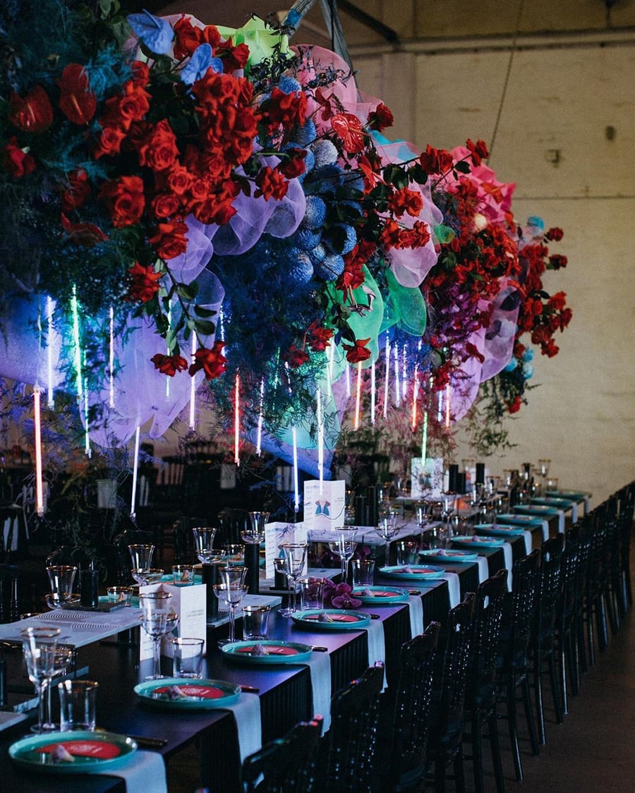 Electric Disco Wedding Ideas |  Modern wedding reception decor featuring a large hanging centrepiece consisting of clusters of red roses, blue pom pom flowers, trailing greenery, clouds of green, pink and purple tulle and vertical neon tubes in red, green and purple that look as if they are floating. This is suspended over a long black table topped with green plates and gold rimmed glassware. | Photography: Varga Murphy via The Wildflower Perth