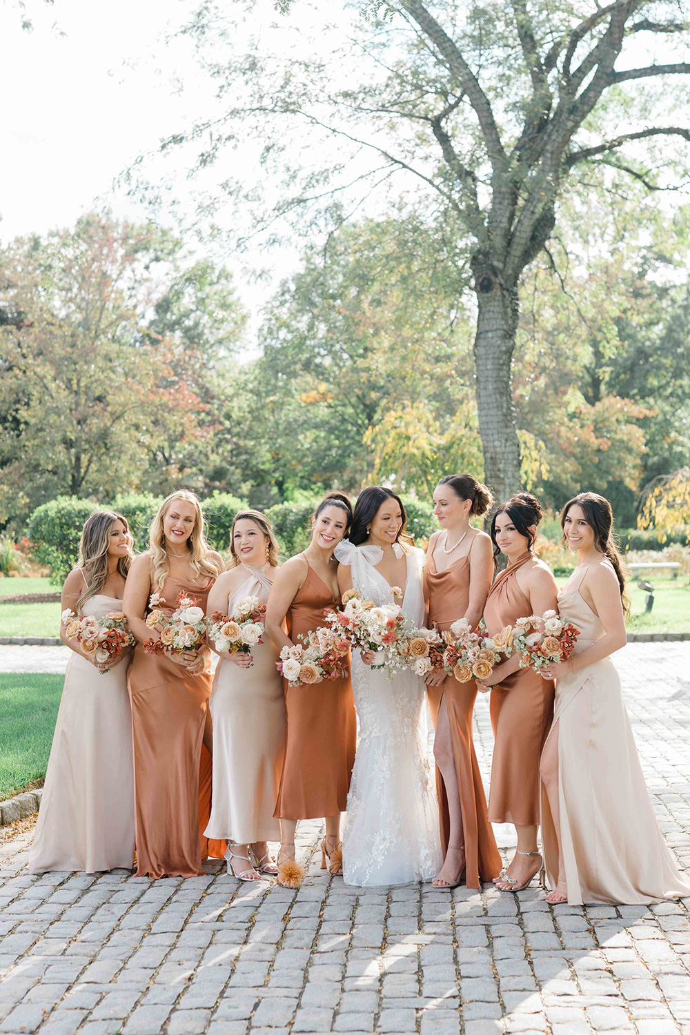 An autumnal wedding party portrait showcases bridesmaids in a tonal palette of champagne and mocha silk dresses, complemented by rose and orchid bouquets. | Karina Mekel Photography via The Budding Florist