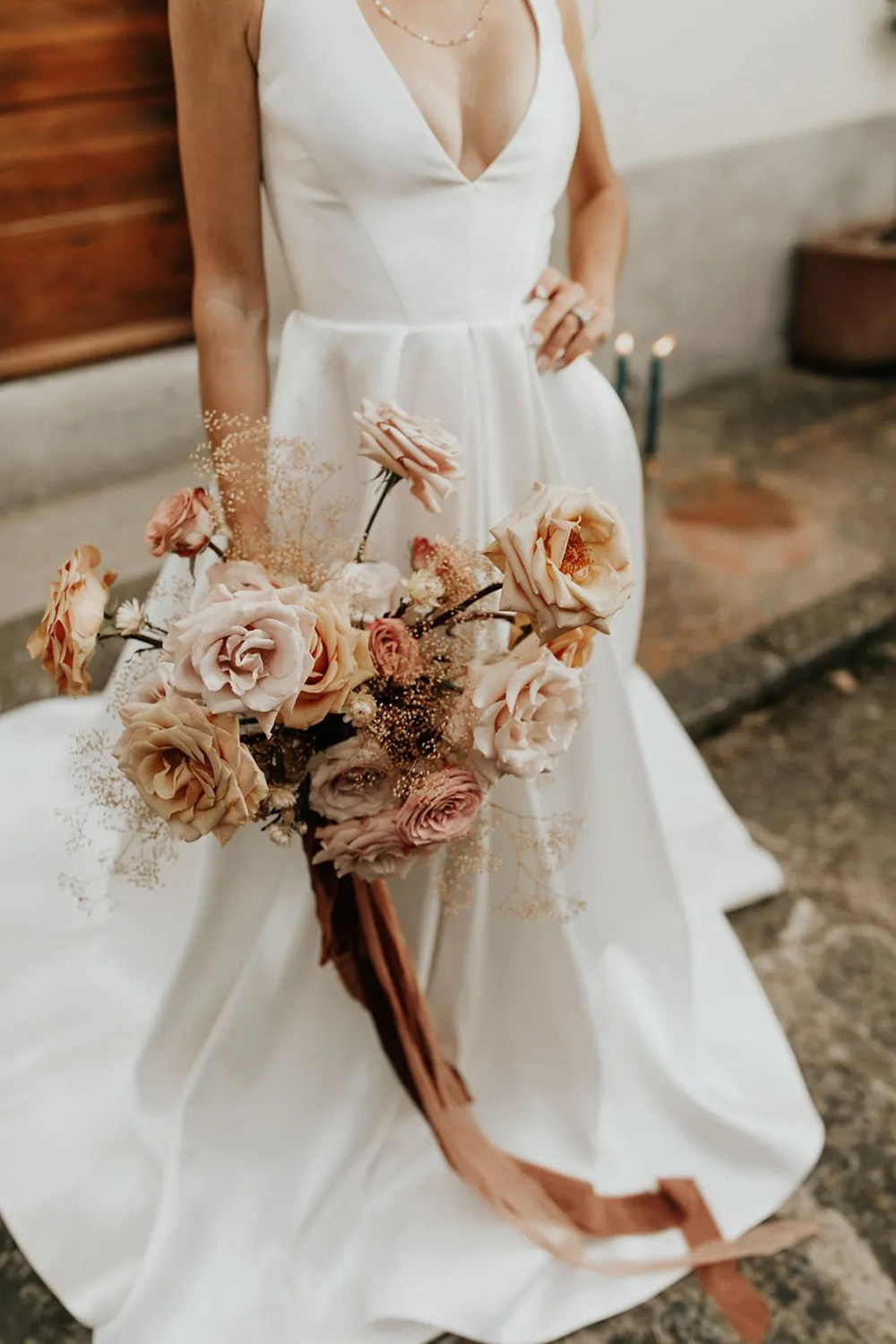 A bride in a modern white satin wedding gown with deep V neckline and fit and flare silhouette holds a romantic bouquet of blush and peach roses with delicate baby’s breath, finished with a cascading mocha-coloured ribbon wrap. | Good Seed Floral