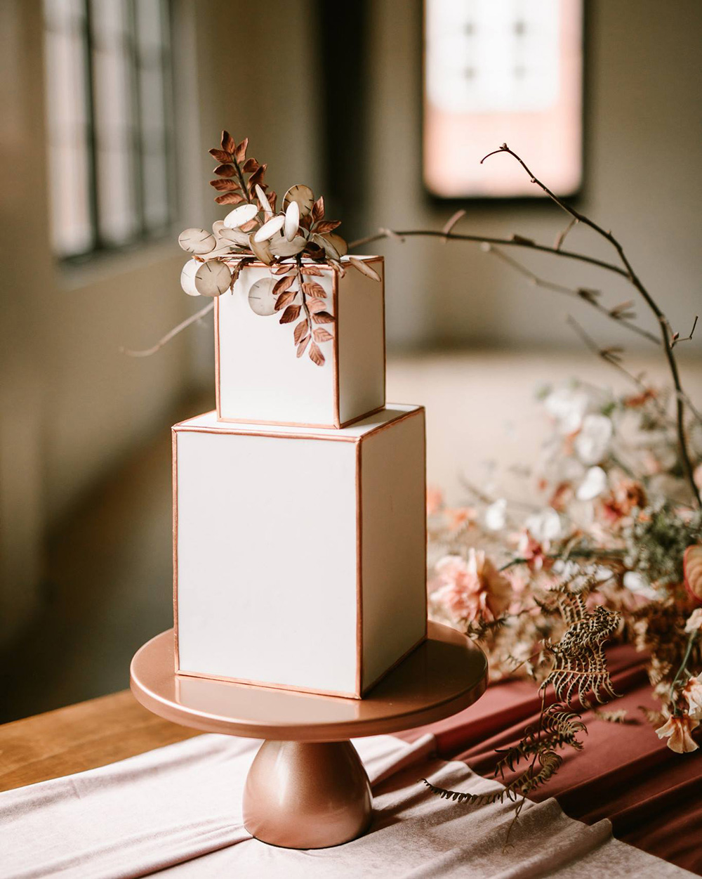 A modern geometric wedding cake in white with copper-toned edges is set on a mocha-coloured cake stand and adorned with metallic foliage accents. | Tiarra Sorte Photographer