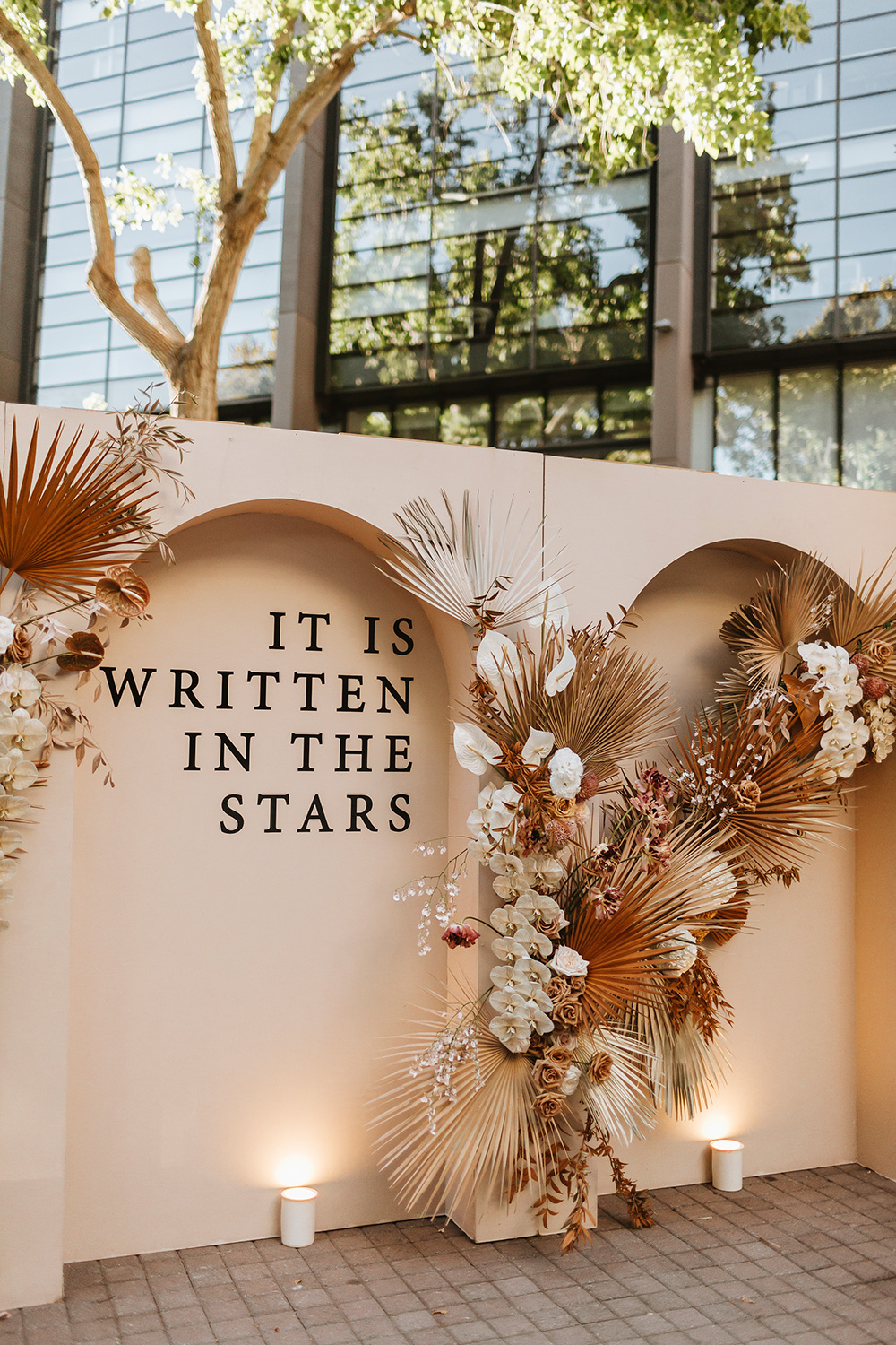 A modern arch wedding backdrop features the romantic phrase, "It is written in the stars" in an elegant serif font, decorated with a dramatic arrangement of boho dried palm fronds, white and champagne orchids, and neutral-toned florals. | Loren Ioppolo via The Lane