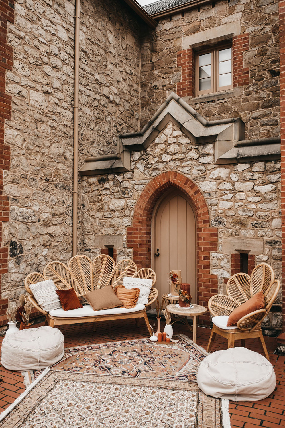 An inviting wedding lounge area features a rattan peacock chair and settee adorned with white cushions and earthy-toned accent pillows, set against an historic stone building. The space is styled with layered vintage-inspired rugs, floor poufs, and small side tables, creating a welcoming bohemian seating area for wedding guests. | Will and Co via Wedshed