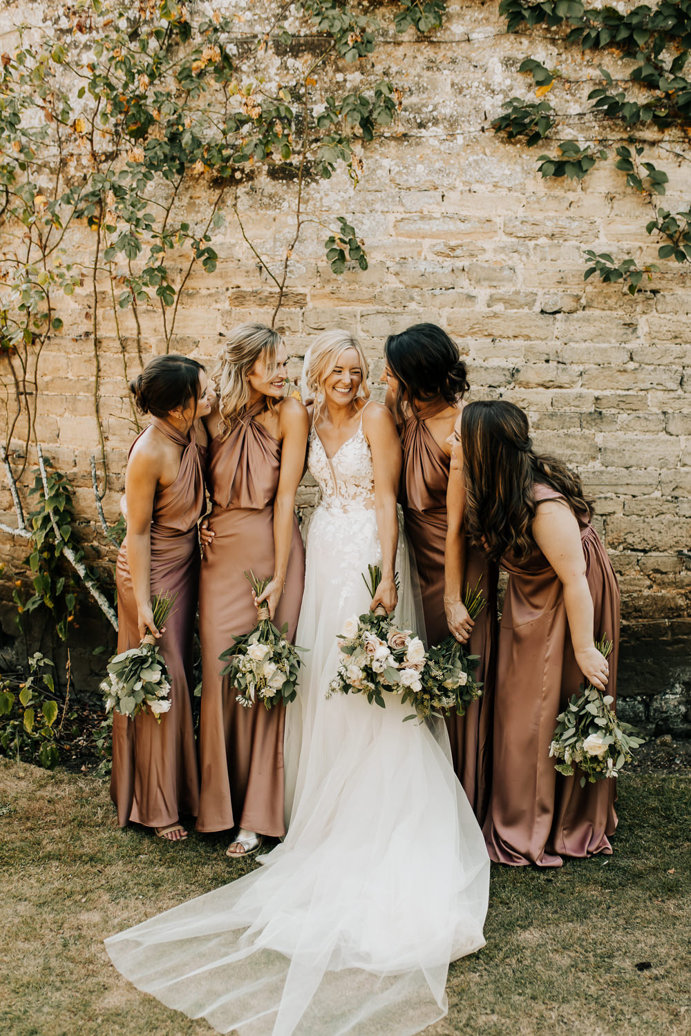 This candid wedding party moment features bridesmaids in mocha-toned silk dresses alongside the bride in a white lace gown, all holding elegant rose bouquets with greenery. | Wyldbee Photography via Whimsical Wonderland Weddings