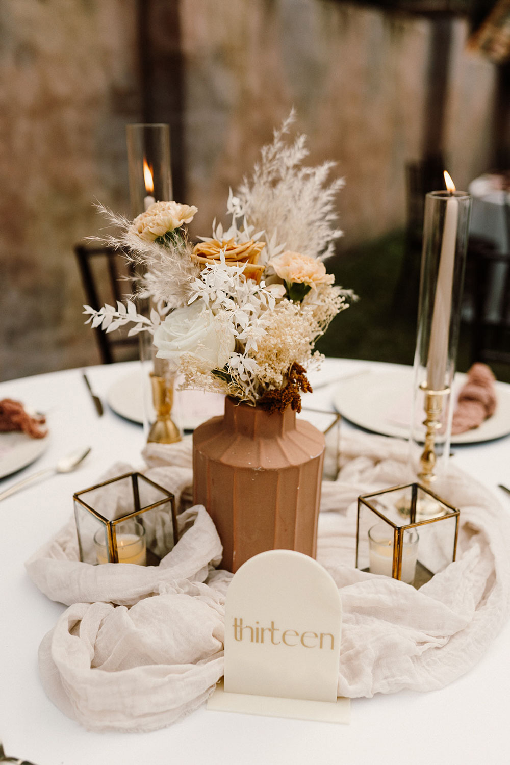 An elegant table centrepiece features dried florals in white and neutral tones displayed in a geometric, mocha-coloured ceramic vase with striped texture. The central arrangement is flanked by brass edged, glass cube lanterns, neutral taper candles in brass candleholders with glass hurricane sleeves, gathered pale muslin fabric and a modern arched acrylic table number with gold lettering. | Josie Brooks Photography via Orange Blossom Bride