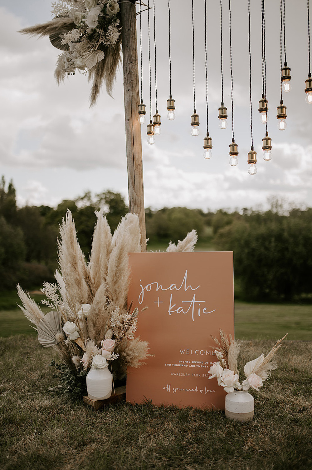 A modern boho welcome sign features a mocha-coloured backdrop with white lettering, Edison bulb lighting installation and pampas grass arrangements in ceramic vases. | Laura Williams Photography via Coco Wedding Venues
