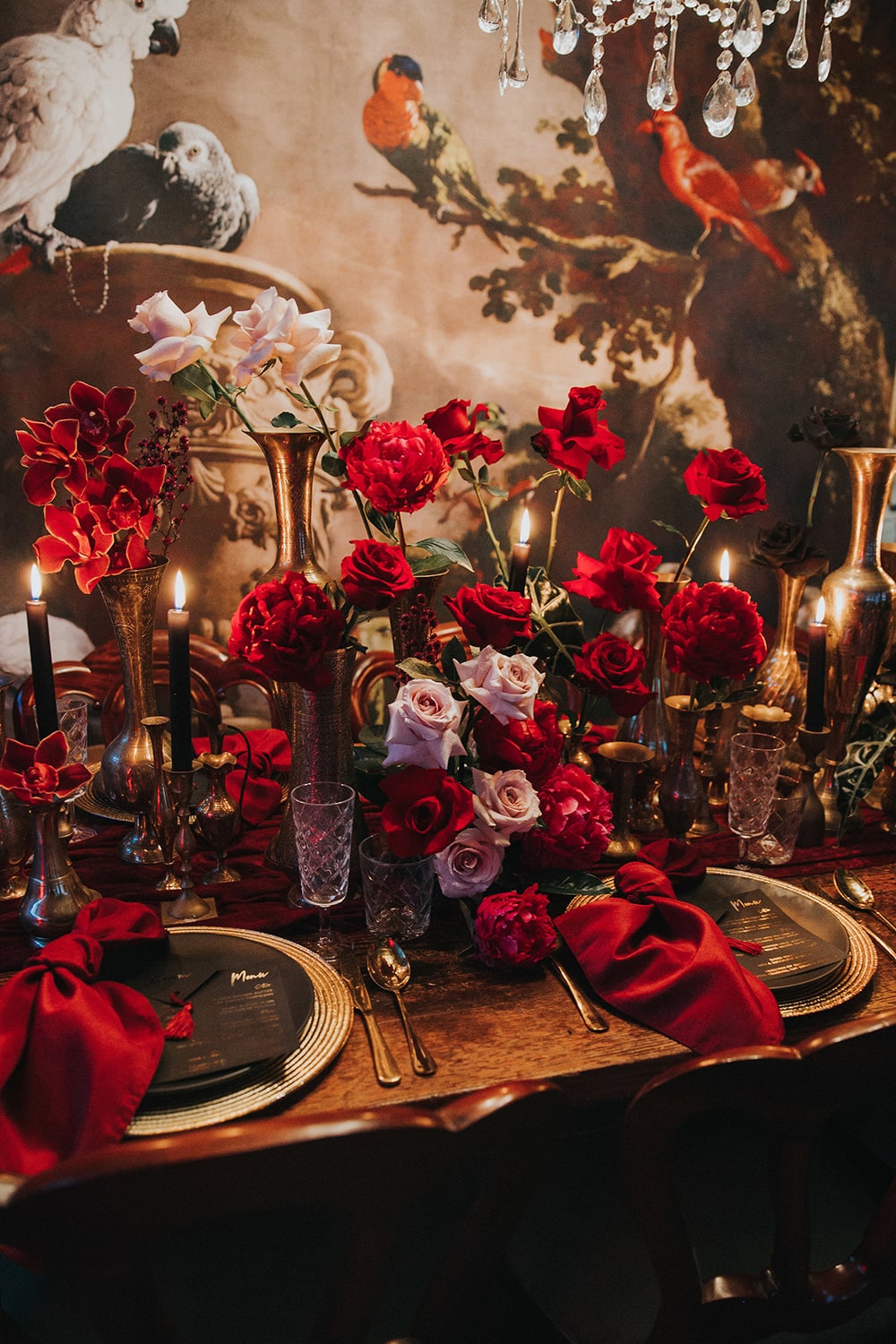 Modern Red & Pink Wedding Ideas | A moody candlelit wedding reception table setting in front of an ornate wall art which features illustrations of exotic birds. The table is decorated with gold vessels of different shapes and sizes filled with clusters of red and pink flowers, gold charger plates and cutlery, black taper candles, plateware and menus, crystal glassware and knotted red napkins. | Photography: White Fox Studios via The Wedding Playbook