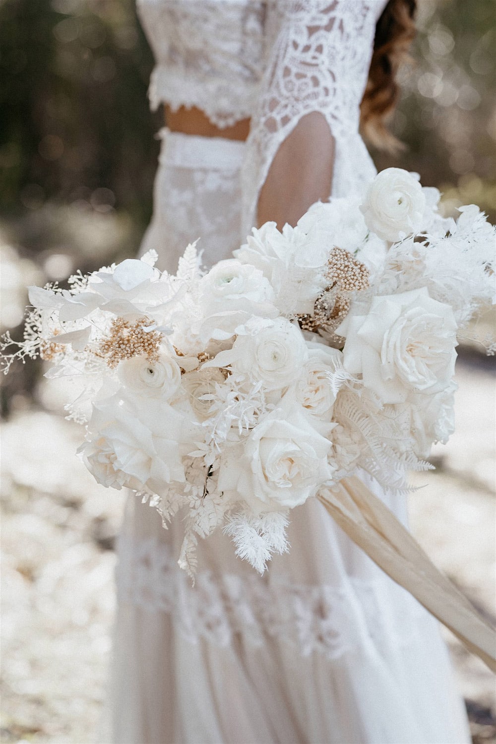 Outdoor Festival Bohemian Wedding Inspiration | This boho bride carries a beautiful white monochromatic wedding bouquet which features ranunculus, roses, fern, silver dollar eucalyptus, ruscus, ming fern, leatherleaf fern, mini gypsophila and neutral rice flower accents tied with champagne ribbon. | Photography: Shae Estella Photo
