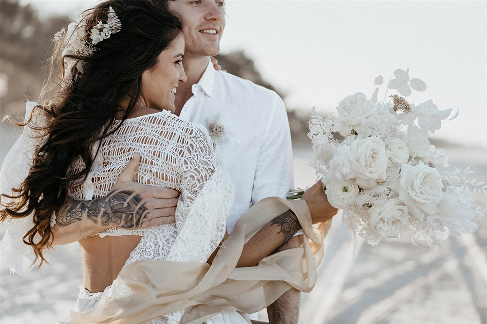 Outdoor Festival Bohemian Wedding Inspiration | The bride wears a white two piece lace boho wedding dress with a long sleeved cropped bodice and tiered maxi skirt. Her hair is worn in a romantic half up half down style with loose waves and a cascading fresh floral hairpiece. The groom wears an untucked white linen shirt with the sleeves rolled up and collar unbuttoned, light grey chinos and brown boots for a casual wedding day look. | Photography: Shae Estella Photo