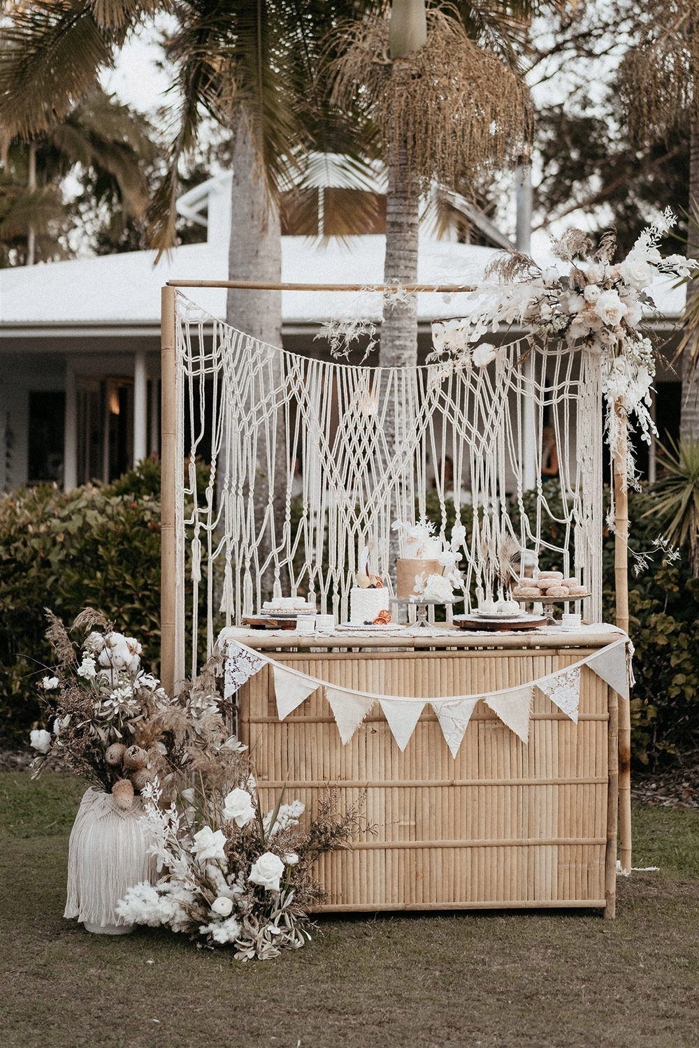 Outdoor Festival Bohemian Wedding Inspiration | For the boho wedding dessert display, a bamboo frame is dressed with an open weave macrame garland and wild neutral and white floral arrangements top right and bottom left. The variety of desserts offers something for everyone, with cakes, donuts, cake pops and macarons presented on a matching bamboo bar decorated with a macrame table runner, white bamboo tealight holders and neutral fabric bunting. | Photography: Shae Estella Photo