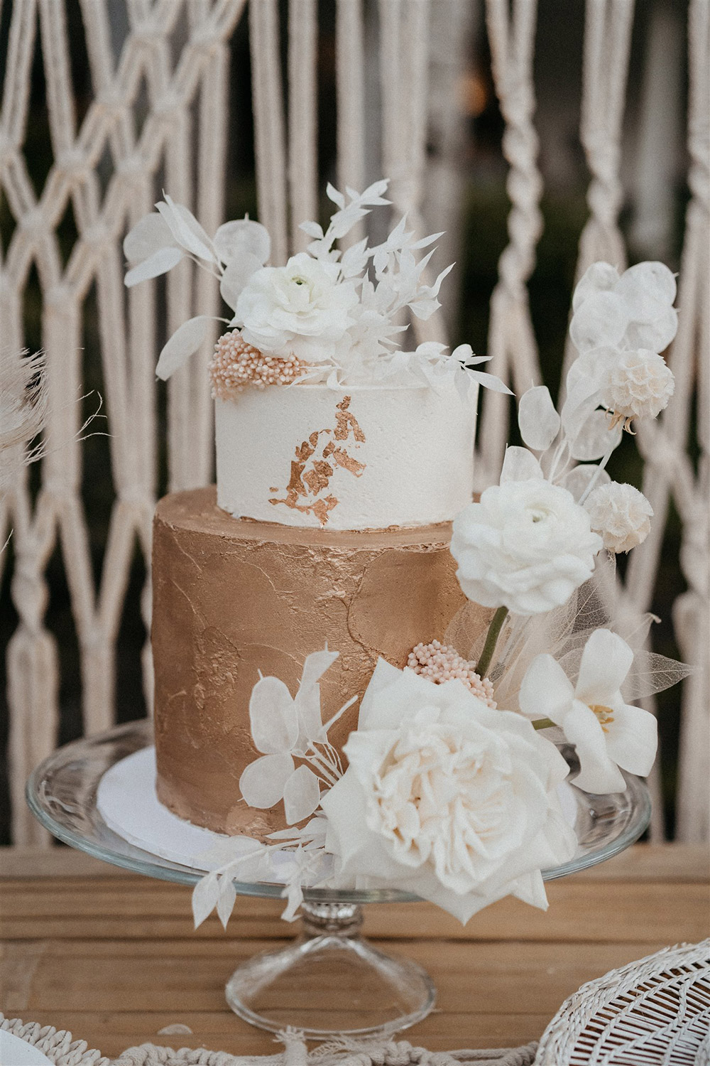 Outdoor Festival Bohemian Wedding Inspiration | A two tiered wedding cake sits on a clear glass cake plate. The top tier features smooth white buttercream with a trail of gold leaf, while the bottom tier features textured gold buttercream. The cake is decorated with artful sprays of white dried silver dollar eucalyptus, ruscus, scabiosa pods and skeleton leaves, blush dried rice flower and fresh white roses, tulips and ranunculus. | Photography: Shae Estella Photo