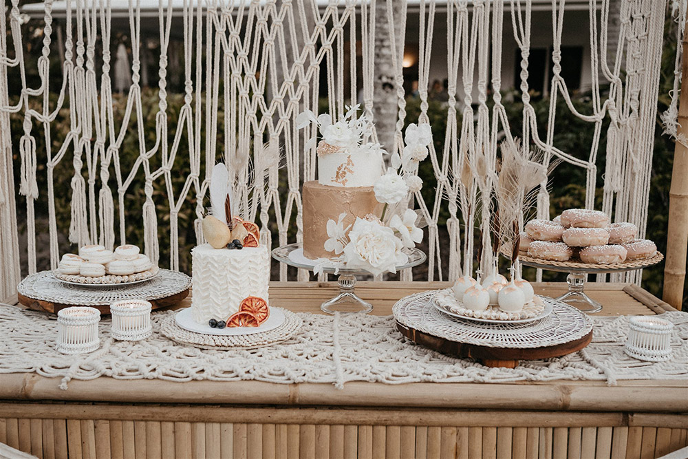 Outdoor Festival Bohemian Wedding Inspiration | For the boho wedding dessert display, a bamboo frame is dressed with an open weave macrame garland and wild neutral and white floral arrangements top right and bottom left. The variety of desserts offers something for everyone, with cakes, donuts, cake pops and macarons presented on a matching bamboo bar decorated with a macrame table runner, white bamboo tealight holders and neutral fabric bunting. | Photography: Shae Estella Photo