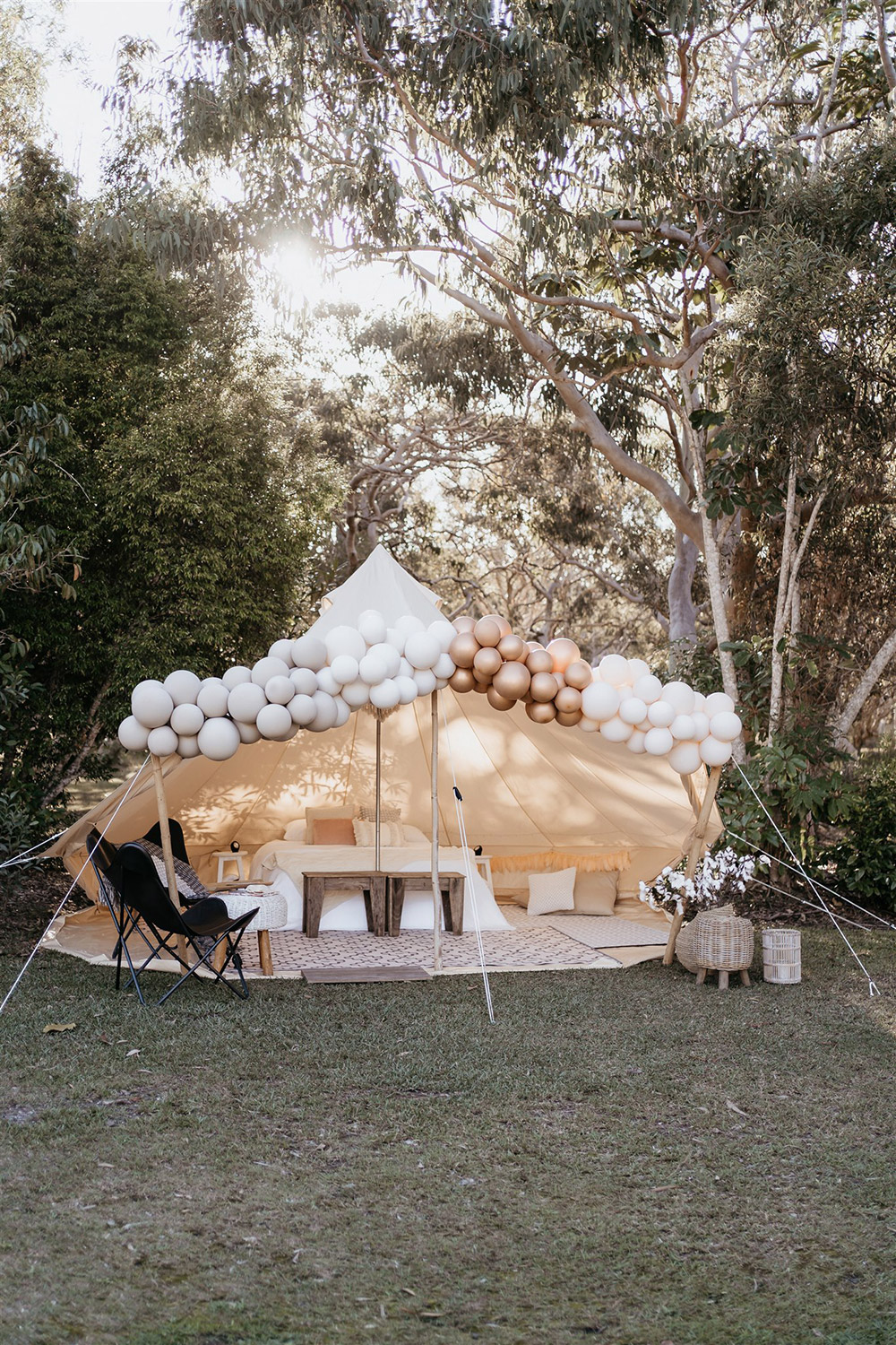 Outdoor Festival Bohemian Wedding Inspiration | A bronze, neutral and white balloon installation frames the entrance to the couple's glamping tent, styled with layered bohemian rugs, throw pillows, a queen bed, camp chairs, fabric garlands, wooden furnishings and wicker decor. | Photography: Shae Estella Photo