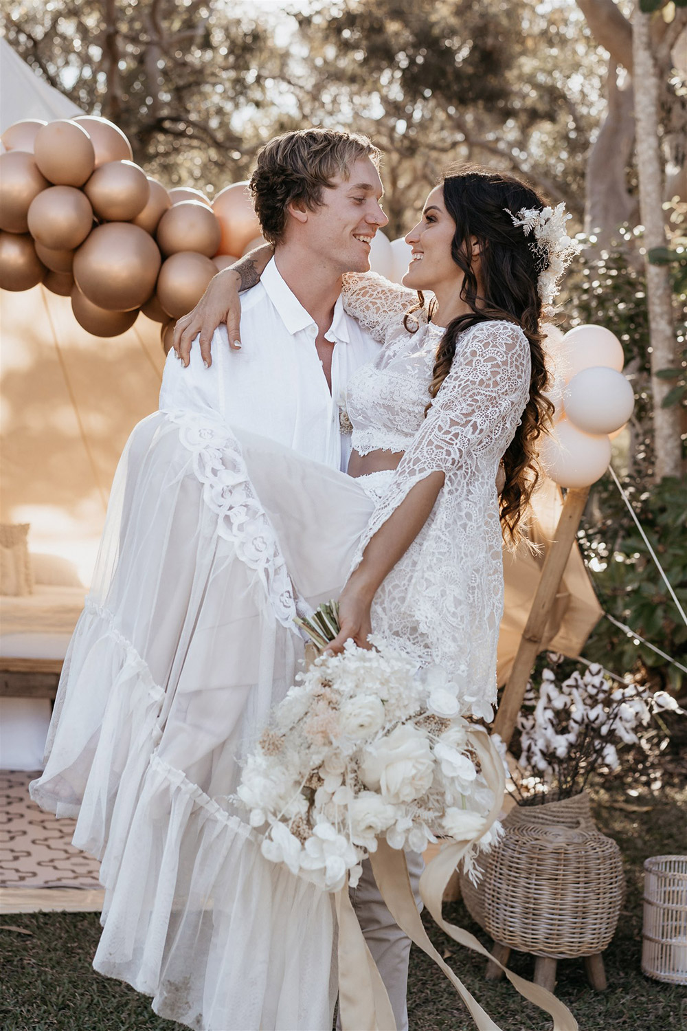 Outdoor Festival Bohemian Wedding Inspiration | A bronze, neutral and white balloon installation frames the entrance to the couple's glamping tent, styled with layered bohemian rugs, throw pillows, a queen bed, camp chairs, fabric garlands, wooden furnishings and wicker decor. | Photography: Shae Estella Photo
