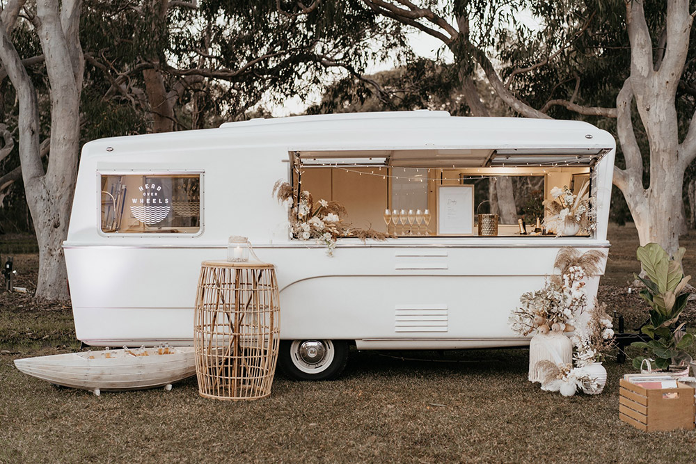 Outdoor Festival Bohemian Wedding Inspiration | After the wedding ceremony, guests are served drinks from a white vintage caravan bar and miniature row boat filled with beer on ice. This setting is styled with white and neutral floral arrangements to match the rest of the boho wedding decor. | Photography: Shae Estella Photo