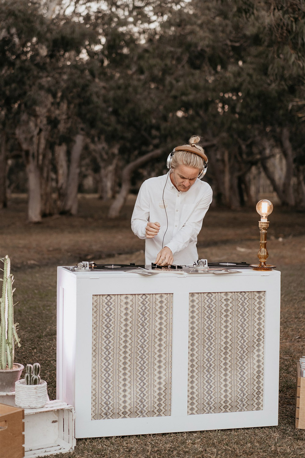 Outdoor Festival Bohemian Wedding Inspiration | A wedding DJ sets a laid-back party atmosphere with the retro sounds of vinyl records. Two neutral geometric fabric panels feature on the front of his white timber DJ station, which is topped with an amber glass table lamp with bare Edison bulb. | Photography: Shae Estella Photo