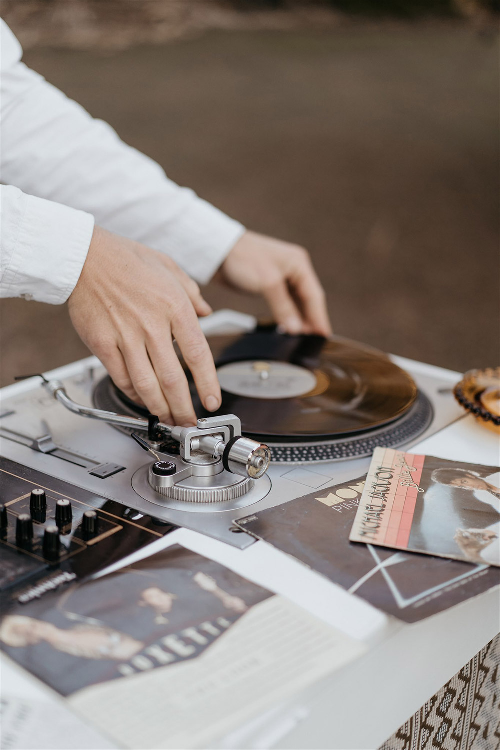 Outdoor Festival Bohemian Wedding Inspiration | A wedding DJ sets a laid-back party atmosphere with the retro sounds of vinyl records. Two neutral geometric fabric panels feature on the front of his white timber DJ station, which is topped with an amber glass table lamp with bare Edison bulb. | Photography: Shae Estella Photo