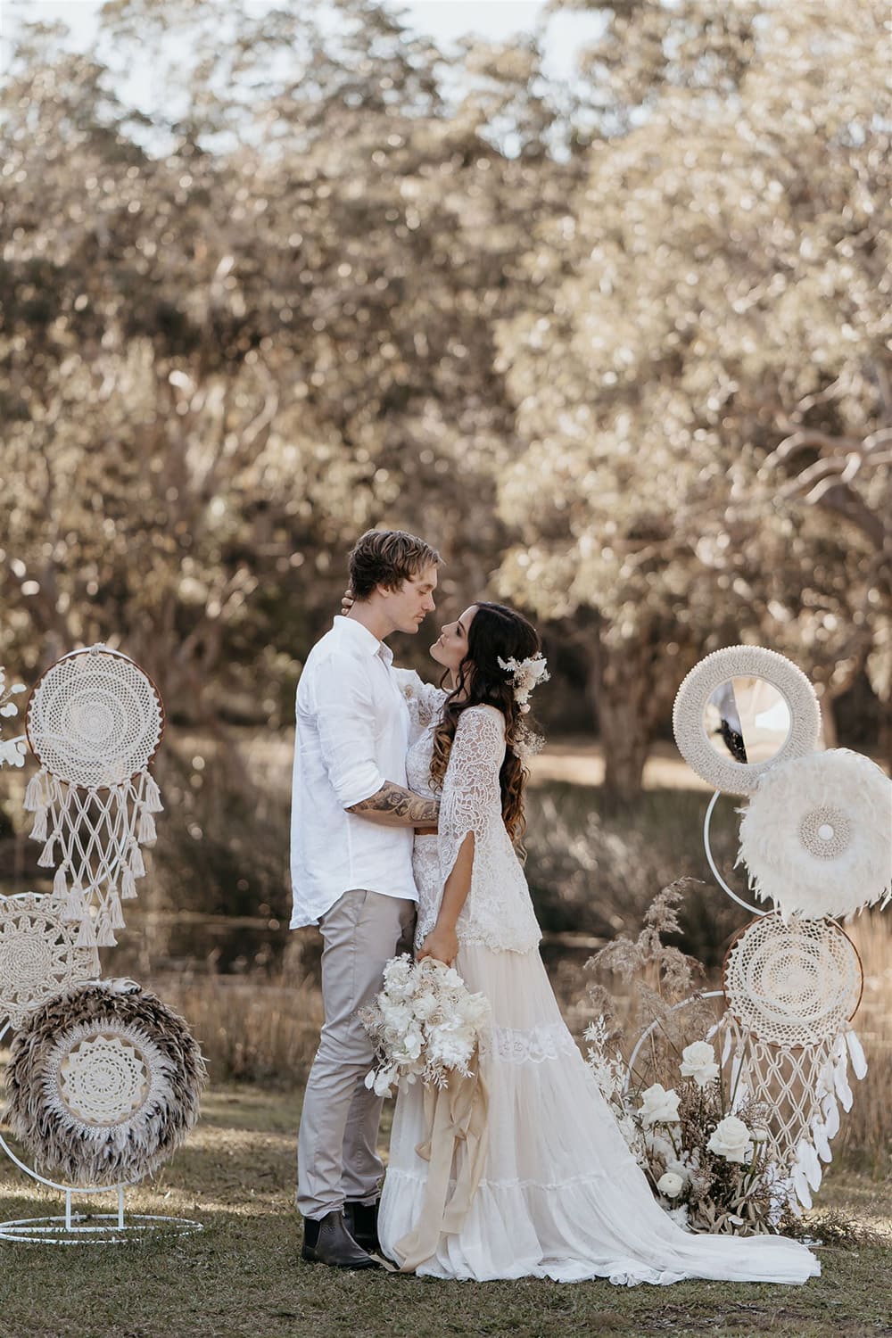 Outdoor Festival Bohemian Wedding Inspiration | This outdoor boho wedding ceremony arbour consists of two pillars of white circles in varying sizes adorned with macrame, feathers and flowers. The bride wears a white two piece lace boho wedding dress with a long sleeved cropped bodice and tiered maxi skirt. Her hair is worn in a romantic half up half down style with loose waves and a cascading fresh floral hairpiece. The groom wears an untucked white linen shirt with the sleeves rolled up and collar unbuttoned, light grey chinos and brown boots for a casual wedding day look. | Photography: Shae Estella Photo