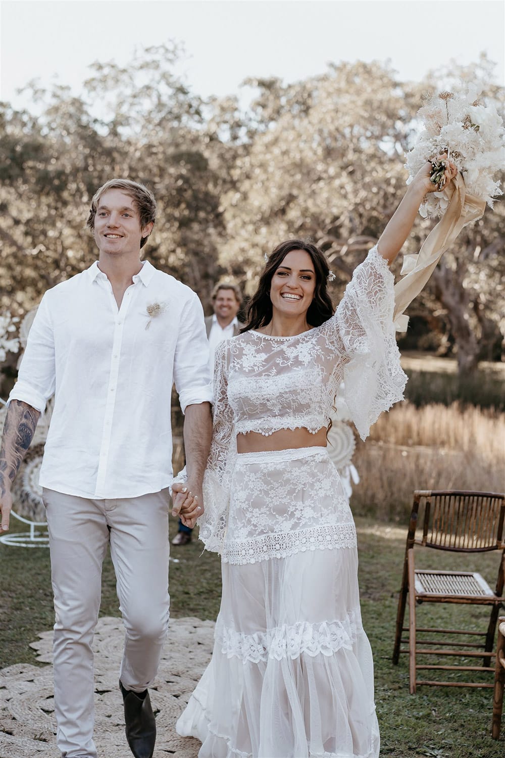 Outdoor Festival Bohemian Wedding Inspiration | The bride wears a white two piece lace boho wedding dress with a long sleeved cropped bodice and tiered maxi skirt. Her hair is worn in a romantic half up half down style with loose waves and a cascading fresh floral hairpiece. The groom wears an untucked white linen shirt with the sleeves rolled up and collar unbuttoned, light grey chinos and brown boots for a casual wedding day look. | Photography: Shae Estella Photo