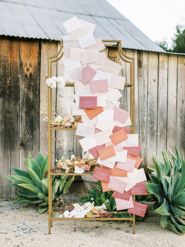 Pantone Peach Fuzz Wedding Ideas | This love letter wedding seating chart features a cascade of envelopes in the colour palette with each guest's name and table handwritten in elegant calligraphy. The envelopes are fixed to a gold shelving display that's styled with matching floral arrangements. | Photography: Ashley Rae Studio