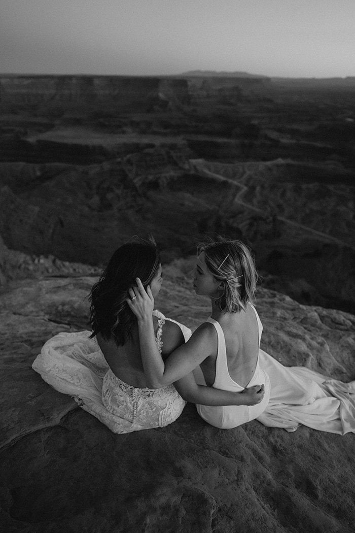 Two brides wearing wedding dresses embrace one another while seated on a clifftop overlooking the mountains. | Photography: Henry Tieu