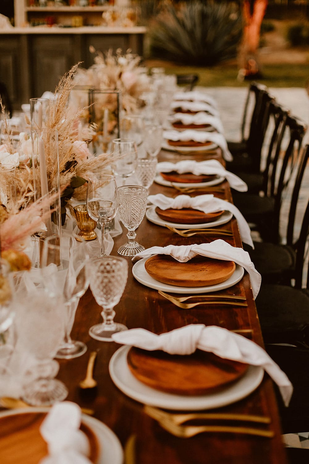 Boho Rust & White Wedding Ideas | A rustic boho wedding reception table setting. The long bare wood table features knotted white napkins atop carved wood plates, paired with gold cutlery, clear embossed glasses, neutral taper candles in clear hurricane vases and low arrangements of roses and pampas grass. The setting is finished with a row of black French provincial inspired cross back chairs. | Photography: Ana & Jerome