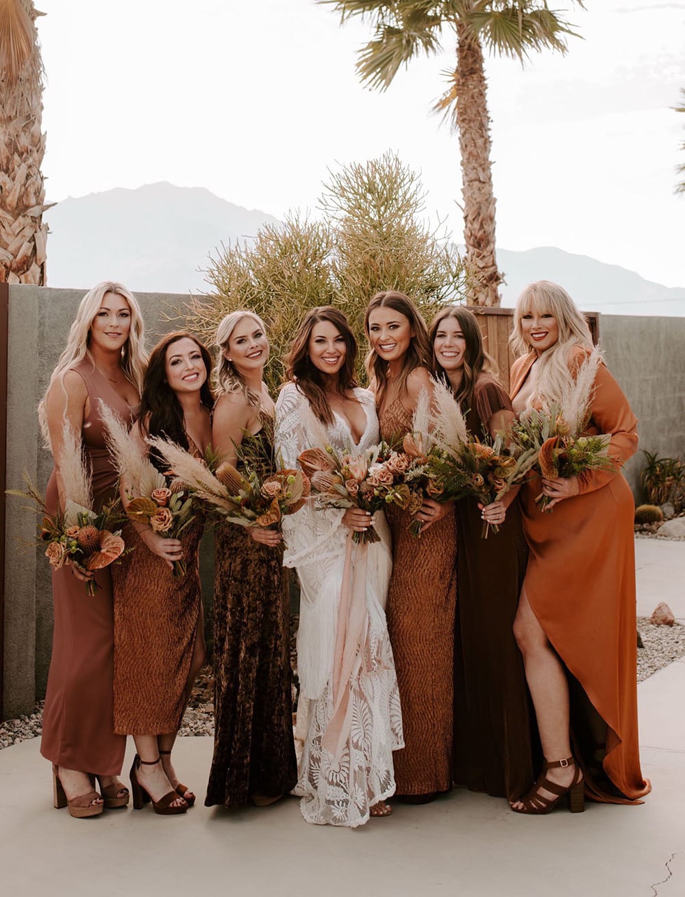 Boho Rust & White Wedding Ideas | Boho bridesmaids wearing mismatched dresses in earthy colours like burnt orange, terracotta and rust. The bride stands in the centre of the group wearing a white boho wedding dress featuring long bell sleeves, a plunging V neckline and lace in a bold foliage pattern. The wedding party wear their hair down in relaxed waves and carry neutral boho wedding bouquets featuring pampas grass, protea, anthurium and roses. The bride's bouquet is tied with long flowing blush ribbons. | Photography: Ashley M. Clayton via Green Wedding Shoes