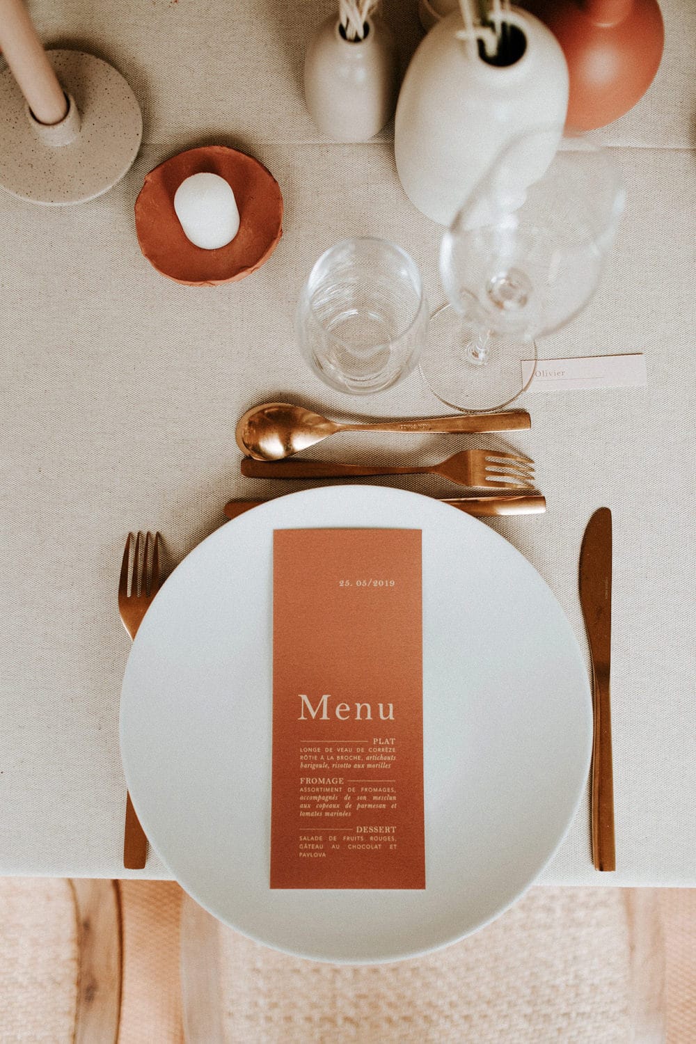 Boho Rust & White Wedding Ideas | A minimalist wedding reception place setting features an oatmeal linen tablecloth, round white plate topped with rectangular rust coloured menu printed with white text, gold cutlery, a terracotta candle plate and small ceramic vases in earthy colours filled with dried flowers. | Photography: Pinewood Weddings via La mariée aux pieds nus