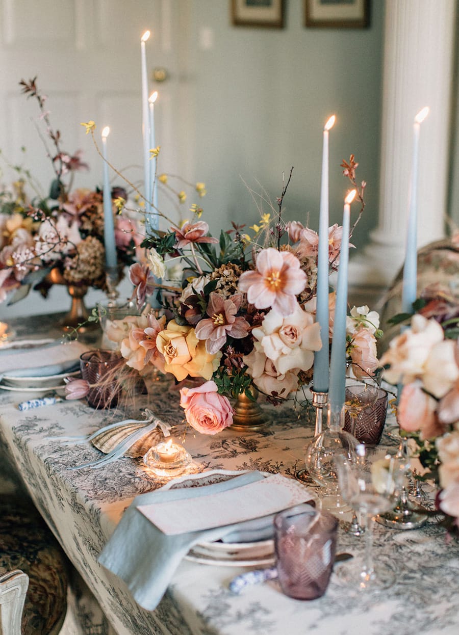 Spring Pastel Wedding Ideas | Romantic wedding reception table setting featuring pale blue taper candles and napkins, purple glassware and rambling floral arrangements in gold footed vases atop a vintage patterned tablecloth. | Photography: Melia Lucida via 100 Layer Cake