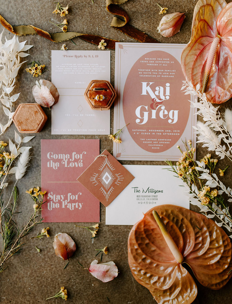 Romantic Boho Terracotta Wedding Ideas | A boho wedding invitation suite in shades of clay, dusty orange and pink terracotta. The invitation features a central coloured oval filled with a white 70s inspired font framed by a double lined border with diamonds in the corners. An accompanying card reads, "Come for the love, stay for the party." A burnt orange hexagon velvet double ring box rests on top of the suite, which is surrounded by dusty orange, blush, yellow and white flowers. | Photography: Ashley M. Clayton via Green Wedding Shoes