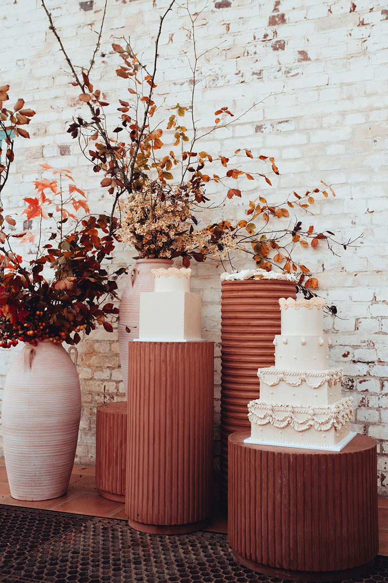 Romantic Boho Terracotta Wedding Ideas | A trio of white wedding cakes sits atop round corrugated terracotta plinths in different heights and diameters alongside tall white ceramic urns filled with autumnal dried foliage in front of a white worn brick wall. | Photography: Chelsea White Photography via Green Wedding Shoes