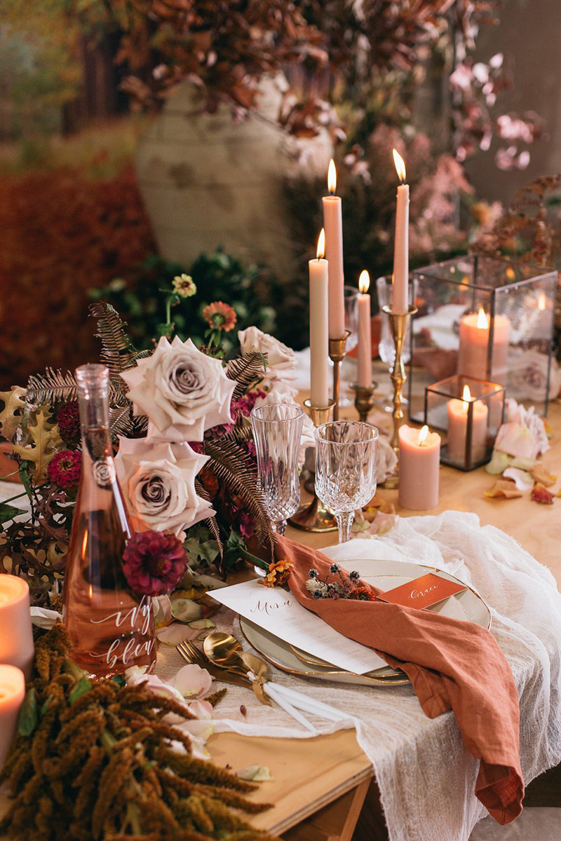 Romantic Boho Terracotta Wedding Ideas | A romantic woodland wedding reception table setting featuring blush candles in vintage brass candlesticks and square glass candle holders, low floral arrangements showcasing pale pink roses, dried foliage and purple blooms, scattered rose petals, vintage crystal stemmed glassware, white and gold dipped cutlery tied with ribbon, and gathered white cheesecloth beneath gold rimmed white ceramic plateware topped with a dusty orange linen napkin, dried flower posy, white menu card and burnt orange place card. | Photography: Figtree Wedding Photography via Ruffled