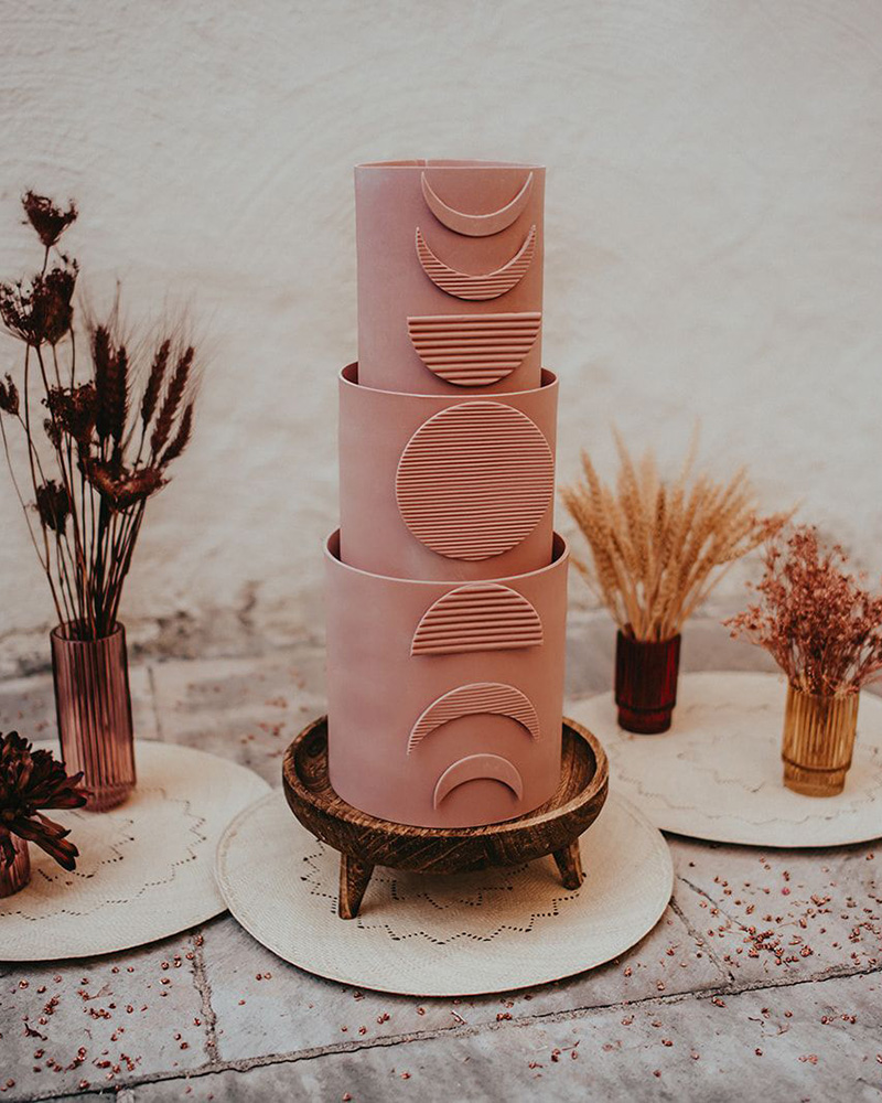 Romantic Boho Terracotta Wedding Ideas | Three tier matte pink terracotta fondant wedding cake featuring moon phases from top to bottom in corrugated texture. The cake is displayed on a carved wooden stand and surrounded by dried flowers in ribbed glass vases. | Photography: Kuenzli Photography via Rock My Wedding