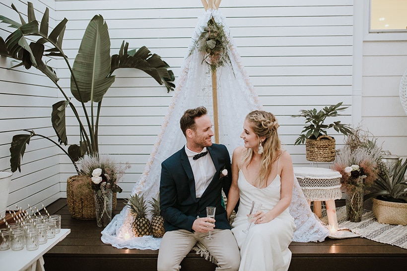 Modern boho bride and groom seated in a lace tipi surrounded by tropical plants for wedding cocktail hour | Alex Jackson Photography