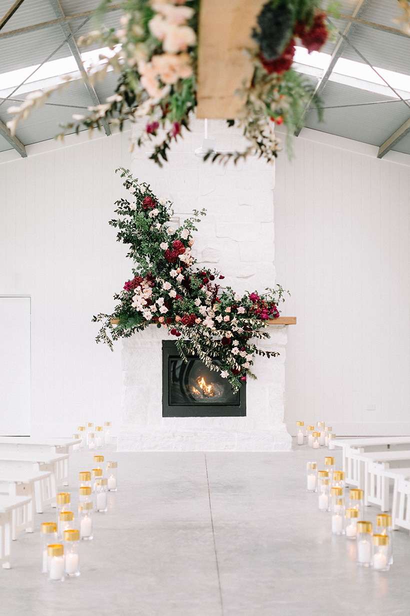 Romantic barn wedding ceremony fireplace backdrop adorned with pink and burgundy flowers at the end of an aisle of white wooden benches and gold pillar candles | Nattnee Photography
