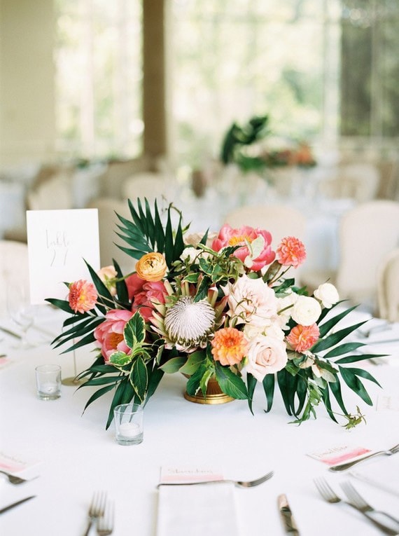 Tropical wedding centrepiece with palm leaves, protea and peony in coral, orange, pink and yellow | Hannah Mayson Photo via 100 Layer Cake